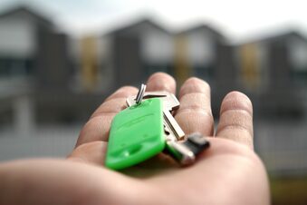 Das Bild zeigt eine Hand, die einen Schlüssel hält. Im Hintergrund ist eine Haus-Silhouette zu sehen