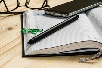 Das Bild zeigt einen Kalender, eine Brille, ein Handy, einen Stift und drei Büroklammern.